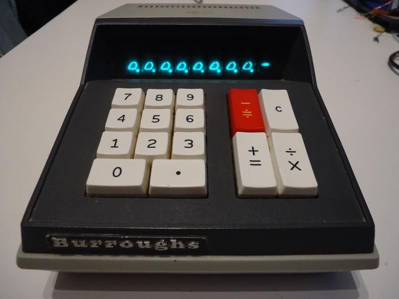 A dark grey calculator with a dark Nixie tube display on top showing zeroes, and a keyboard of thick plastic keys.