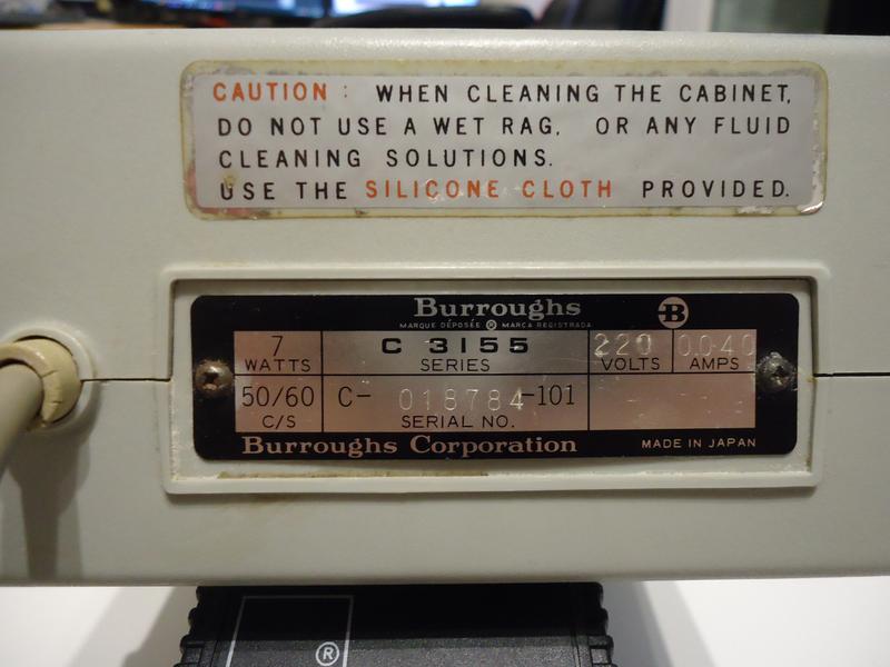 The back of the calculator, with a serial number sticker, and a cleaning information sticker, with the power cable coming out on the left. The cleaning sticker says "Caution: When cleaning the cabinet, do not use a wet rag, or any fluid cleaning solutions. Use the silicone cloth provided.". The serial number sticker says (left to right, top to bottom) "Burroughs, 7 Watts, C3155 Series, 220 Volts, 0.040 Amps, 50/60 C/S, C-018784-101 Serial No. Made in Japan".