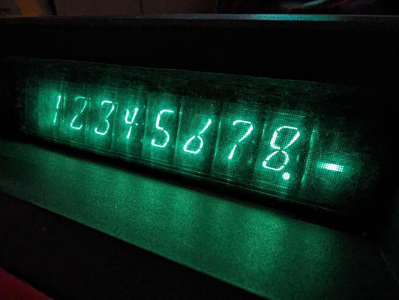 A close-up of the nine Nixie tubes, showing the digits 1 to 8, followed by a decimal point and a minus sign. The environment is dark. The tubes glow a bright green tint.
